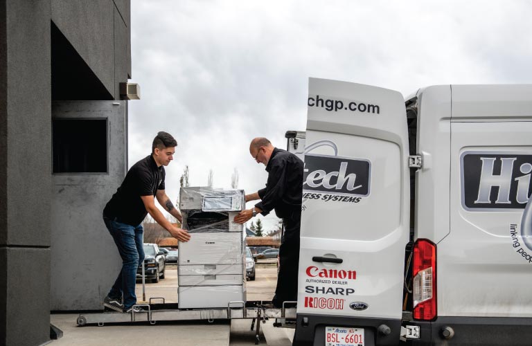 Two employees load a multi-functional printer (MFP) into a company delivery van